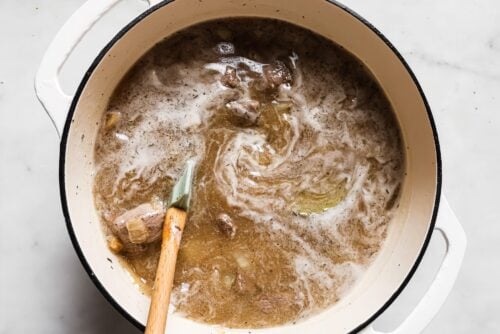 A white Dutch oven filled with simmering Apple Cider Beef Stew, showing chunks of meat, a bay leaf, and bubbling broth, being stirred with a wooden spoon on a marble countertop.