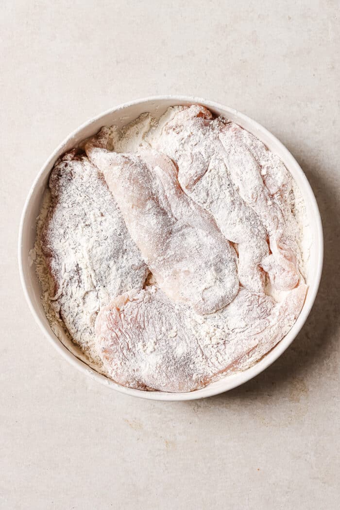 A large white bowl full of chicken cutlets dredged in flour. 