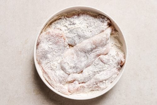 Raw chicken breast fillets, soon to become Honey Garlic Chicken Breast, are coated in flour and arranged in a white bowl on a light-colored surface, ready for cooking.