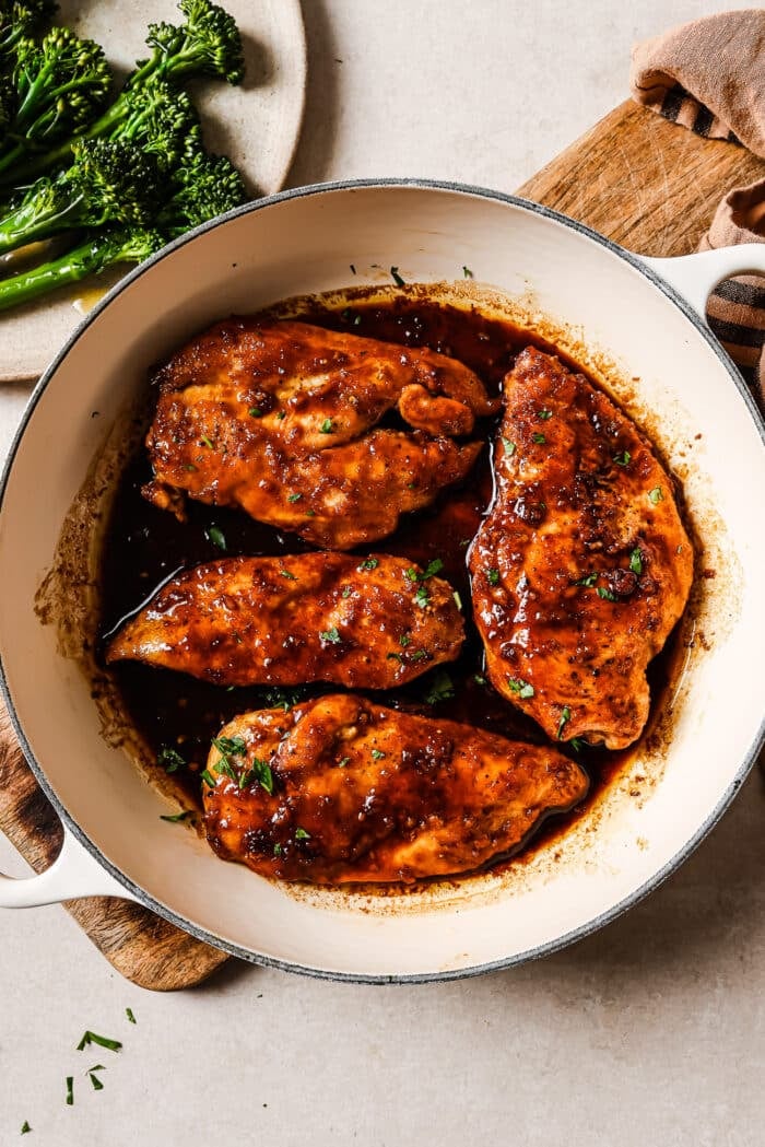 Honey garlic chicken breast garnished with fresh parsley in a white skillet with broccoli on the side. 