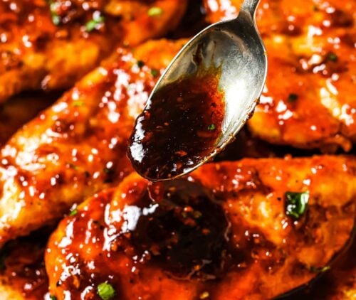 A spoon drizzles thick, glossy honey garlic barbecue sauce over golden-brown, cooked chicken breasts in a skillet, garnished with fresh herbs.