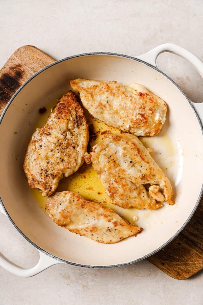 An overhead view of four seared chicken cutlets in a white skillet. 