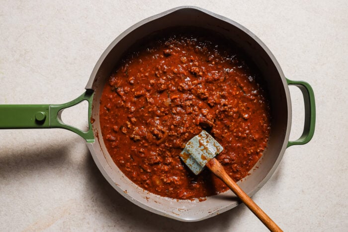 A green-handled skillet filled with thick, chunky Baked Ziti with Ground Beef sauce sits on a light-colored surface. A wooden spatula with a blue head rests in the savory mixture.