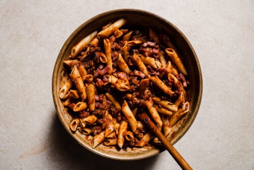 A bowl of penne pasta mixed with a chunky tomato-based meat sauce and bits of bacon, reminiscent of Baked Ziti with Ground Beef, with a wooden spoon resting inside the bowl on a light-colored surface.