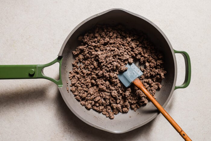 A green-handled skillet filled with cooked ground beef for Baked Ziti with Ground Beef and a blue spatula rests on a light-colored countertop.