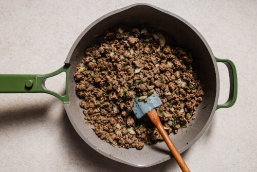 A green-handled skillet filled with cooked ground beef, chopped onions, and herbs—perfect for making Baked Ziti with Ground Beef. A spatula with a wooden handle and blue blade rests in the pan, which sits on a light-colored surface.