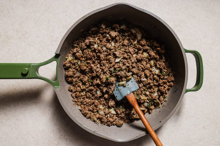 A green-handled skillet filled with cooked ground beef, chopped onions, and herbs—perfect for making Baked Ziti with Ground Beef. A spatula with a wooden handle and blue blade rests in the pan, which sits on a light-colored surface.