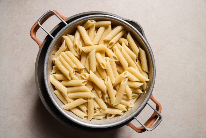 A metal colander filled with cooked penne pasta, perfect for making Baked Ziti with Ground Beef, sits on top of a pot. The background features a light-colored countertop.