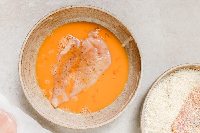 A raw chicken breast sits in a shallow bowl filled with beaten eggs and black pepper, ready to become crispy Air Fryer Chicken Cutlets. Nearby are a plate of breadcrumbs and another piece of raw chicken on a light-colored surface.