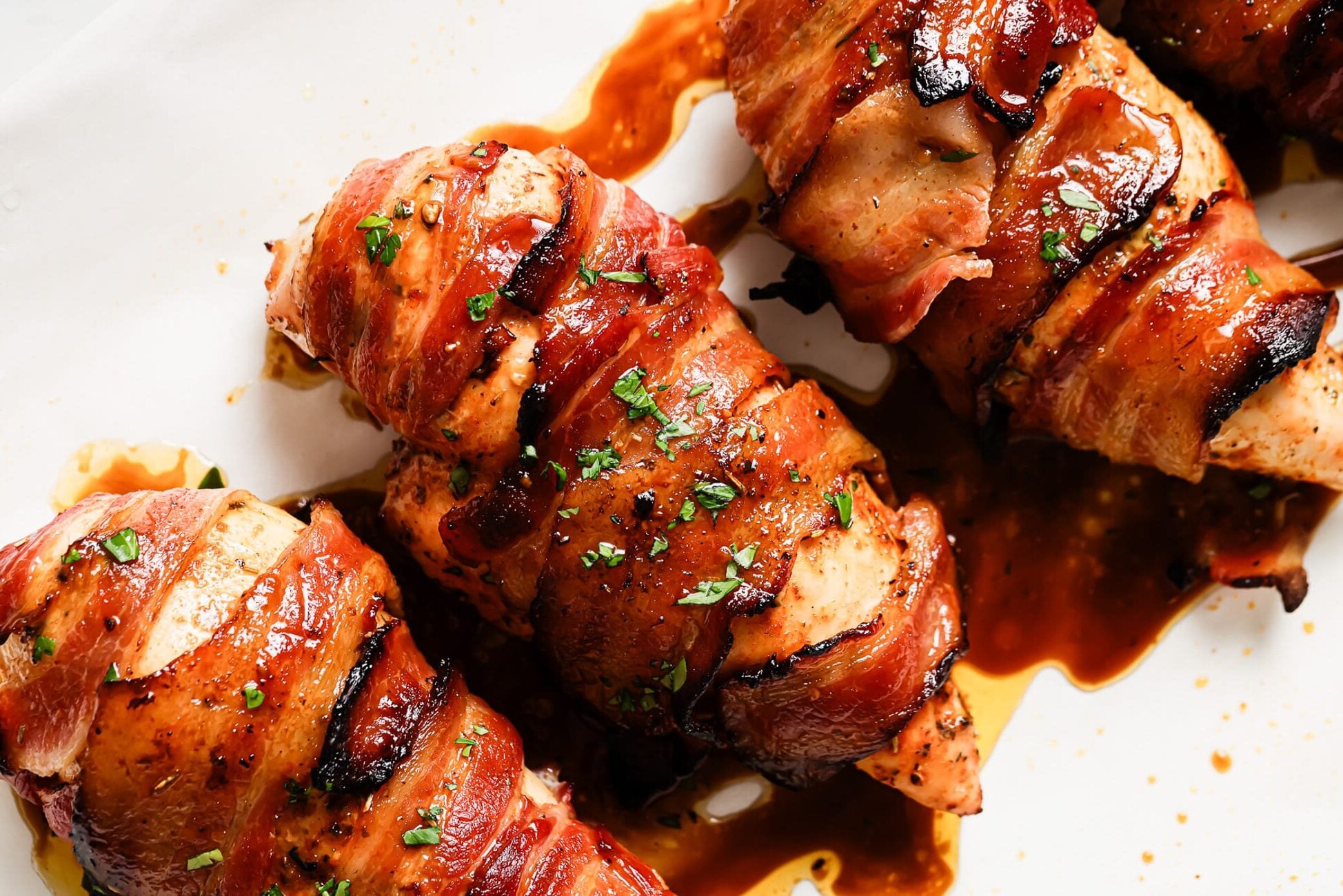 Close-up of Bacon Wrapped Chicken Breast covered in a glossy brown sauce, garnished with chopped parsley, on a white plate.