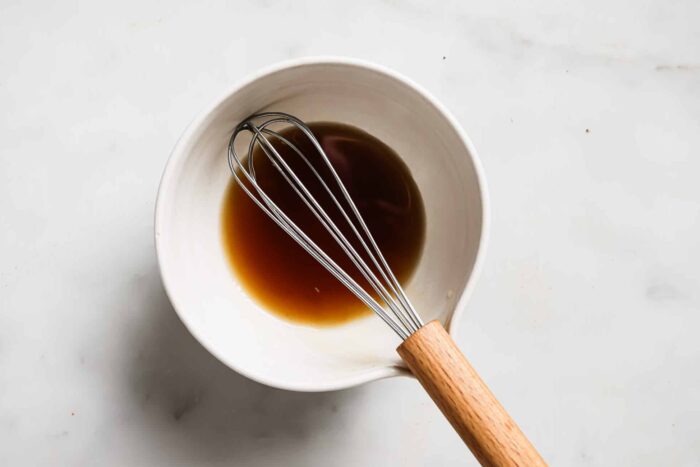 A metal whisk with a wooden handle rests in a white bowl containing a small amount of brown liquid, perhaps the perfect marinade for Bacon Wrapped Chicken Breast, all placed on a light-colored surface.