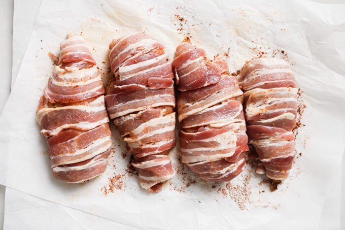 Four Bacon Wrapped Chicken Breasts are arranged on white parchment paper, each wrapped in strips of bacon with a sprinkle of seasoning visible.