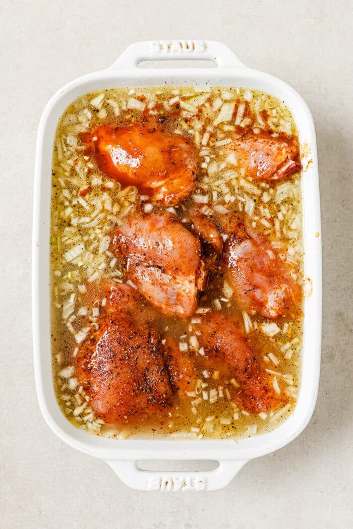 Six marinated chicken thighs in a white casserole dish with uncooked rice, garlic, and onions.