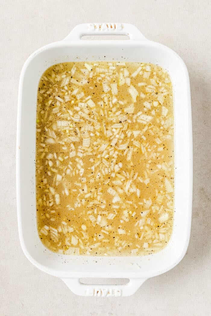 A white casserole dish filled with garlic, onions, oil, butter, hot water, rice, and seasonings.