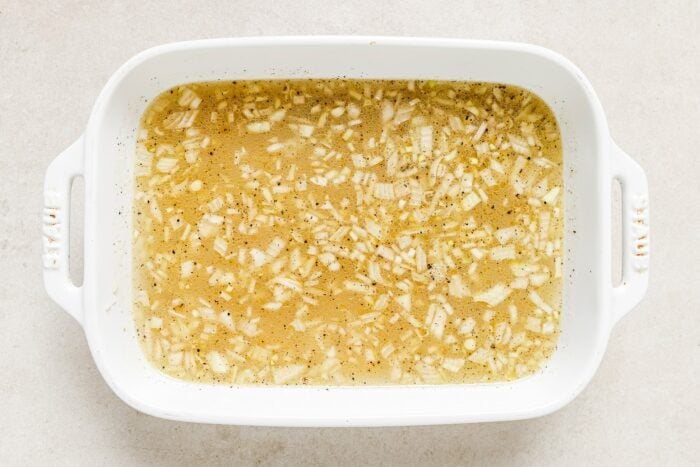 A white rectangular baking dish filled with broth, finely chopped onions, black pepper, and a layer of Baked Chicken Thighs and Rice rests on a light-colored surface.