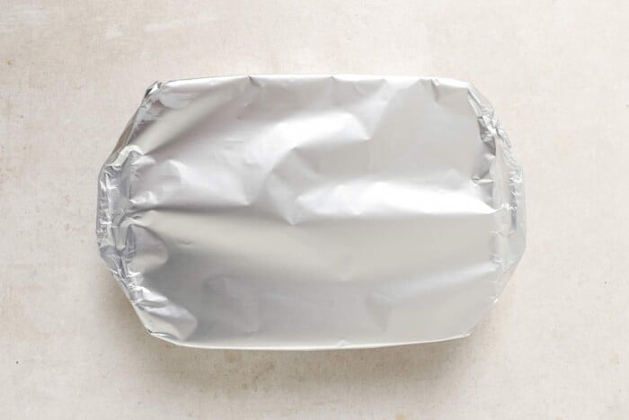 A rectangular baking dish, perfect for Baked Chicken Thighs and Rice, is covered tightly with a sheet of aluminum foil and set on a light-colored surface.