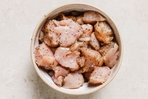 A bowl filled with raw, diced chicken pieces seasoned for Garlic Butter Chicken Bites with herbs and spices, placed on a light-colored surface.