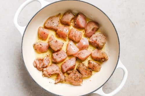 Raw, seasoned chicken breast pieces in a white Dutch oven, coated with oil and herbs for delicious Garlic Butter Chicken Bites, ready to be cooked. The pot is placed on a light-colored surface.