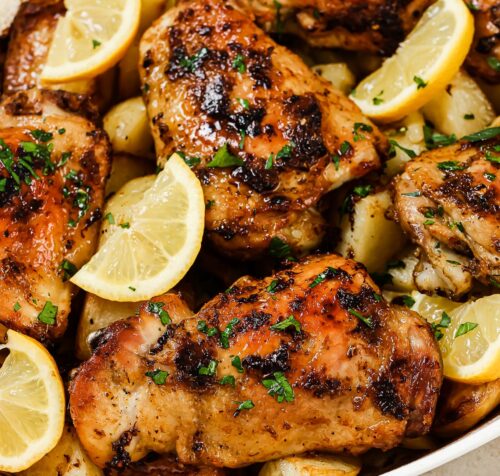 Greek Lemon Chicken and Potatoes featuring roasted chicken thighs with golden crispy skin, garnished with chopped herbs and served alongside roasted potatoes and lemon wedges in a white dish.