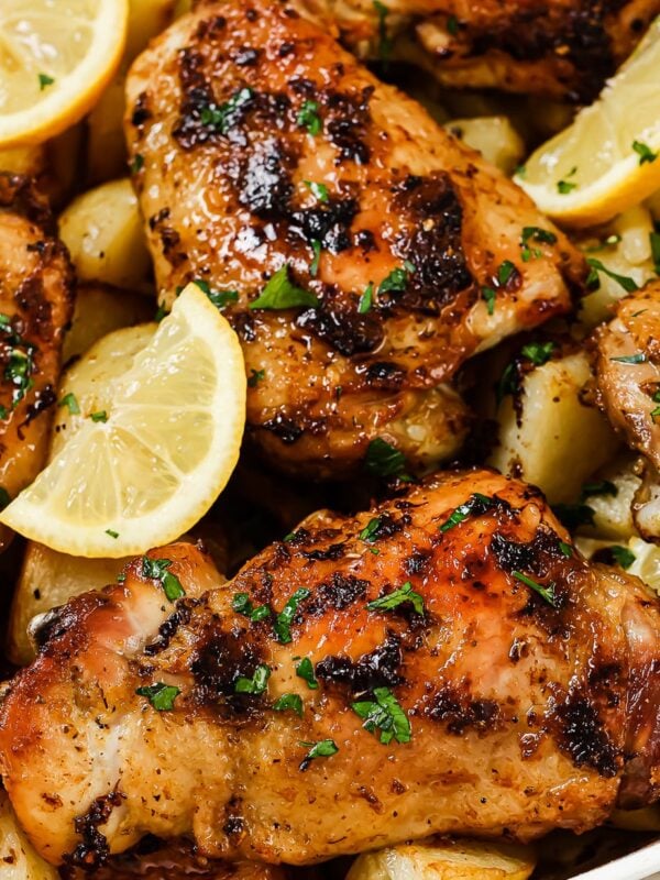 Greek Lemon Chicken and Potatoes featuring roasted chicken thighs with golden crispy skin, garnished with chopped herbs and served alongside roasted potatoes and lemon wedges in a white dish.
