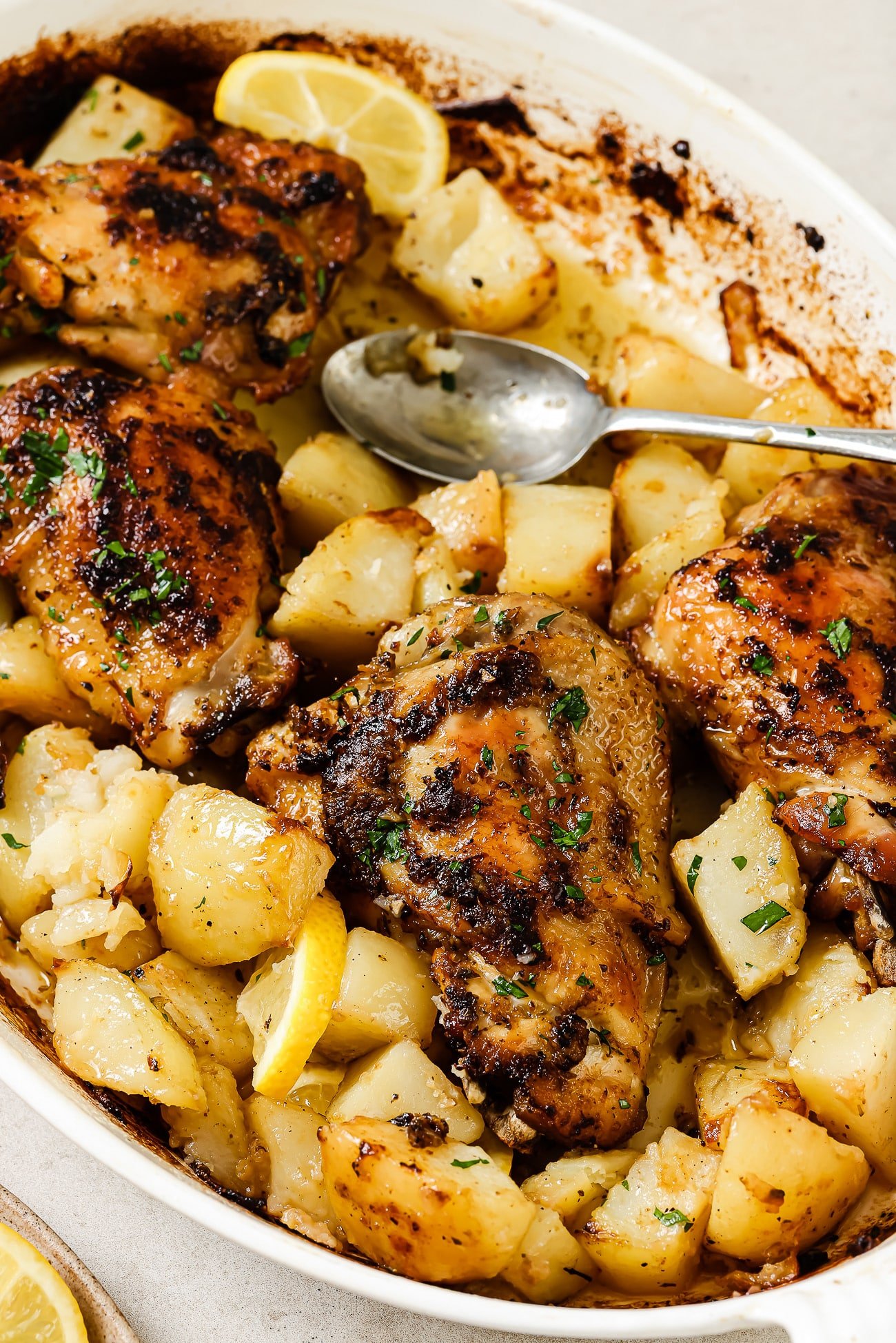 Baked Greek chicken and potatoes in a white baking dish with a serving spoon in it.