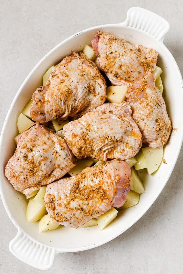 An overhead view of marinated chicken thighs over diced potatoes in a white baking dish.