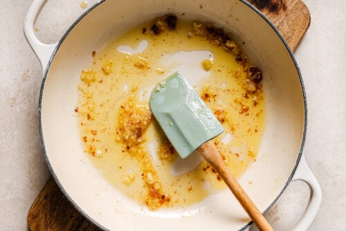 A white enameled pot contains sautéed minced garlic and oil, with browned bits on the bottom—the perfect base for Honey Garlic Chicken Breast. A light green spatula with a wooden handle rests inside the pot, which sits on a wooden trivet.