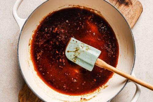 A pale blue spatula stirs a reddish-brown Honey Garlic Chicken Breast sauce with visible minced garlic in a white enameled pot, which is sitting on a wooden cutting board and a light-colored countertop.