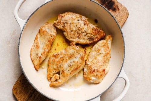 Four golden-brown Honey Garlic Chicken Breasts in a white, round pan with handles, resting in a small amount of oil on a wooden board, against a light background.