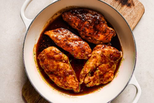 Four cooked Honey Garlic Chicken Breasts are covered in a glossy, dark brown sauce, resting in a white Dutch oven on a wooden surface.