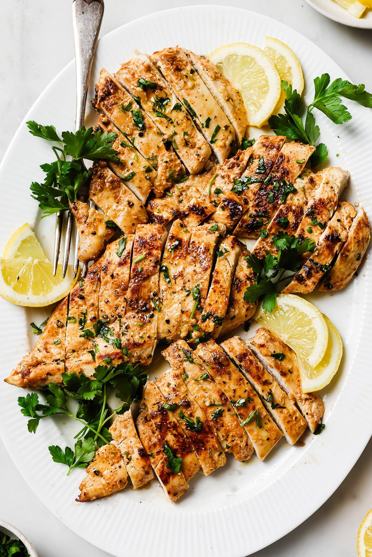 Yogurt-marinated chicken breast sliced and garnished with parsley and lemon wedges on a white serving plate. 