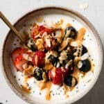 A bowl of yogurt and cottage cheese topped with strawberries, raspberries, blackberries, blueberries, chia seeds, almond slices, and a drizzle of nut butter, with a spoon inside—perfect for a wholesome Cottage Cheese Breakfast Bowl.