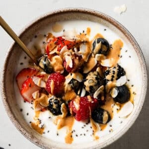 A bowl of yogurt and cottage cheese topped with strawberries, raspberries, blackberries, blueberries, chia seeds, almond slices, and a drizzle of nut butter, with a spoon inside—perfect for a wholesome Cottage Cheese Breakfast Bowl.