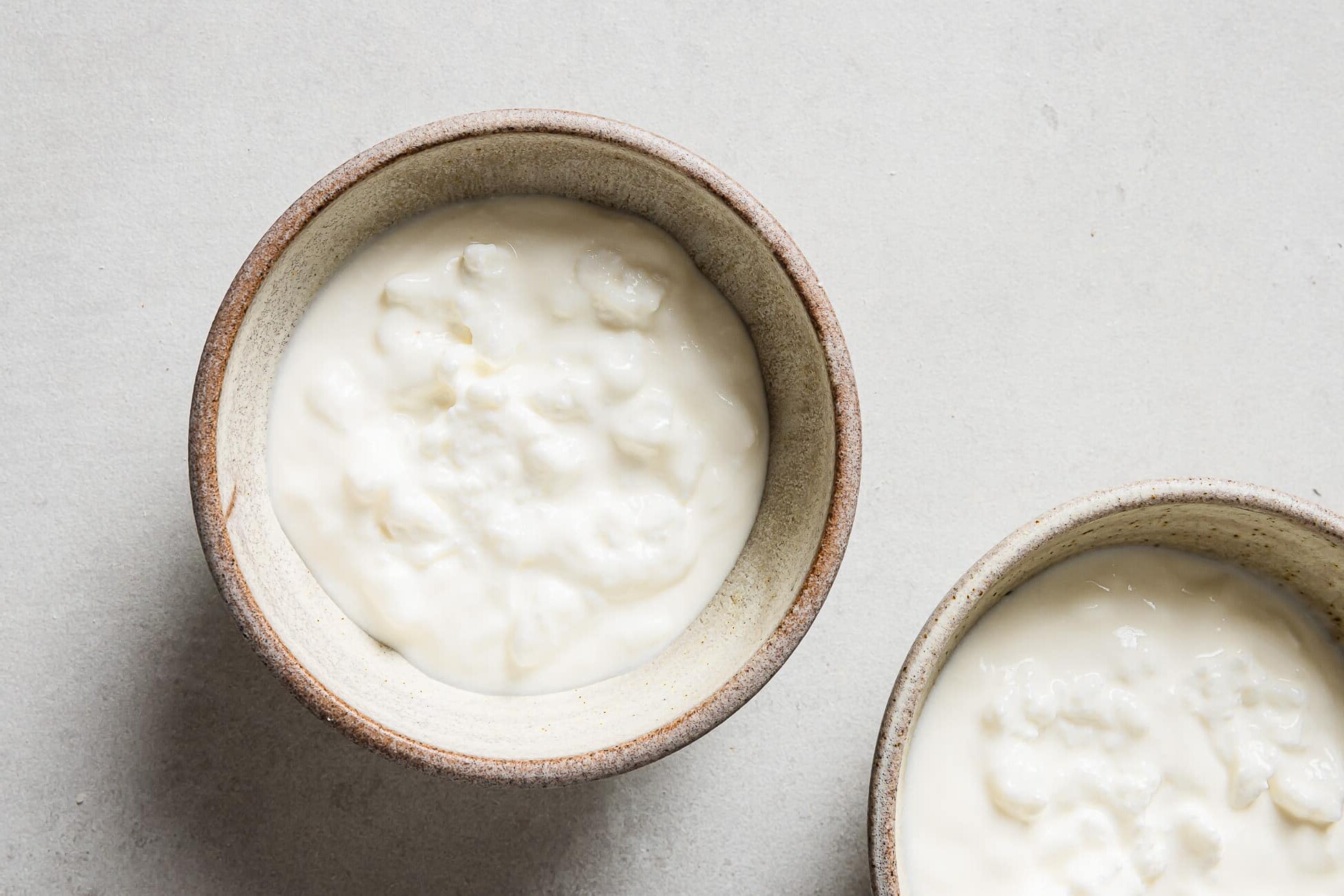 Two ceramic bowls, each filled with creamy, white yogurt with a slightly lumpy texture reminiscent of a Cottage Cheese Breakfast Bowl, sit on a light gray surface. Only part of the second bowl is visible in the lower right corner.