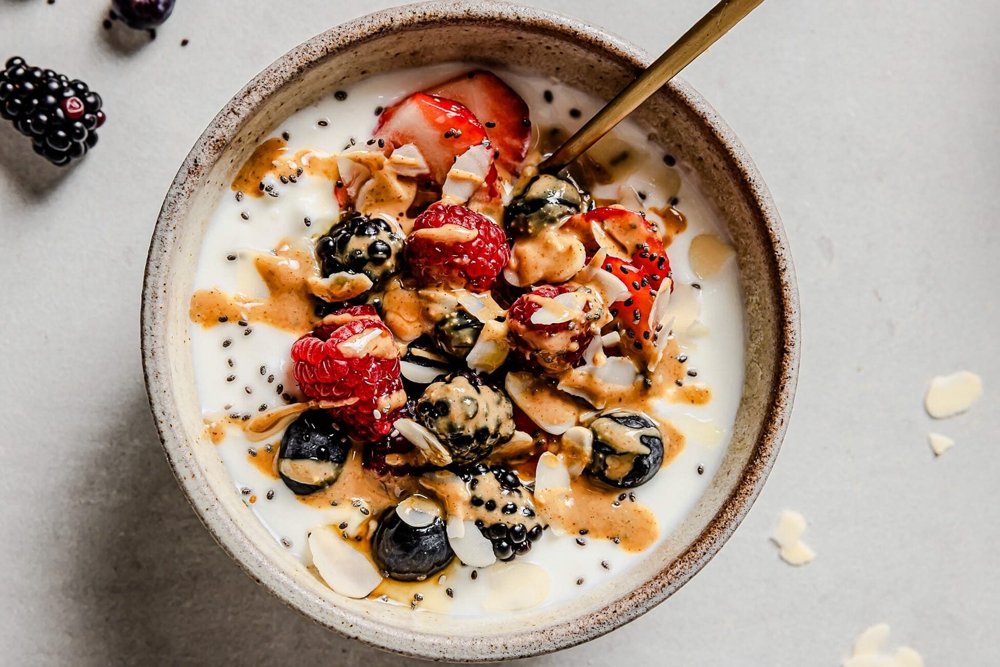 A delicious Cottage Cheese Breakfast Bowl sits ready to enjoy, topped with strawberries, raspberries, blueberries, blackberries, chia seeds, sliced almonds, and a drizzle of nut butter. A spoon rests in the bowl while a few berries are scattered on the surface nearby.