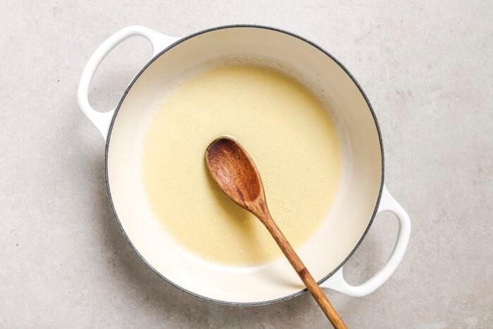 A white pot with handles contains a light yellow sauce or roux, being stirred with a wooden spoon, perfect as the base for Garlic Butter Chicken Bites, on a light-colored surface.
