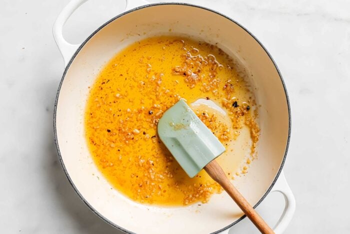 A white pot on a marble surface contains sautéed minced garlic in oil, perfect for starting garlic butter chicken tenders, being stirred with a mint green spatula.