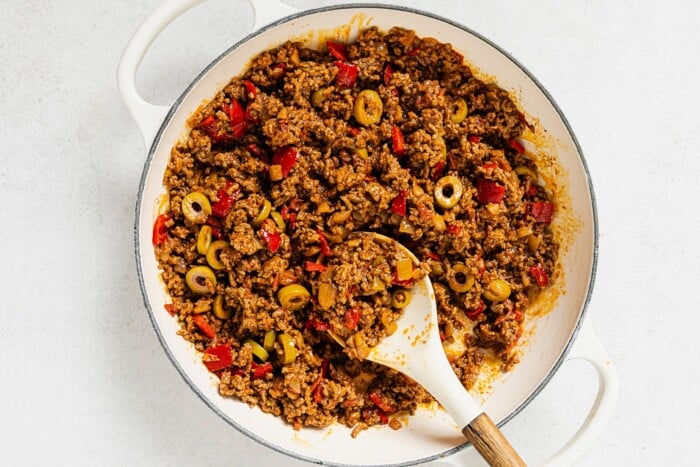 A white skillet filled with cooked ground beef mixed with red bell peppers and sliced green olives, showcasing a classic Picadillo Recipe, with a wooden spoon resting in the savory mixture.