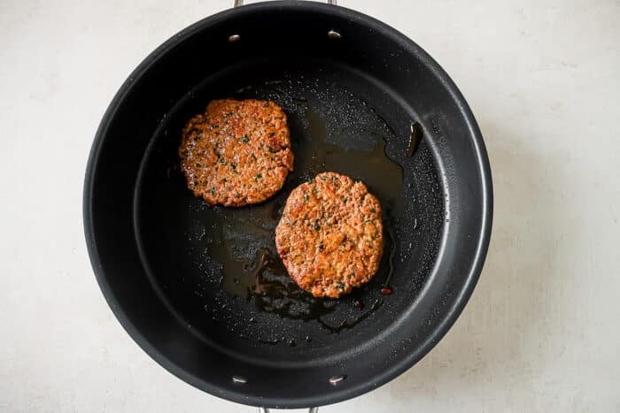 Two ground turkey burger patties cooking in a large skillet.