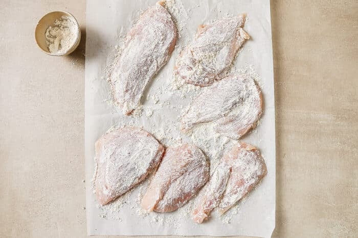 Six chicken cutlets coated in salt, pepper, and flour on a sheet of parchment paper.