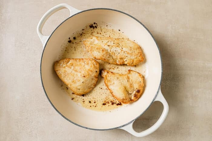 Three chicken cutlets coated in flour are seared in a cast-iron Dutch oven.