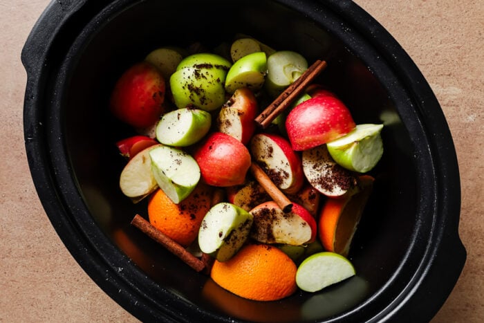 Slow cooker filled with apples, orange slices, cinnamon sticks, and water for homemade apple cider.