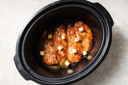 Raw seasoned chicken breasts with cubes of butter on top inside a slow cooker.