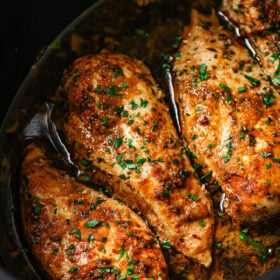 Three seasoned Slow Cooker Chicken Breasts sit in a dark slow cooker with flavorful sauce and sprinkled fresh herbs, showcasing a juicy and tender appearance.