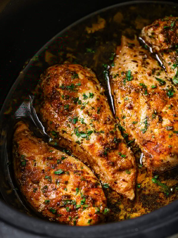 Three seasoned Slow Cooker Chicken Breasts sit in a dark slow cooker with flavorful sauce and sprinkled fresh herbs, showcasing a juicy and tender appearance.