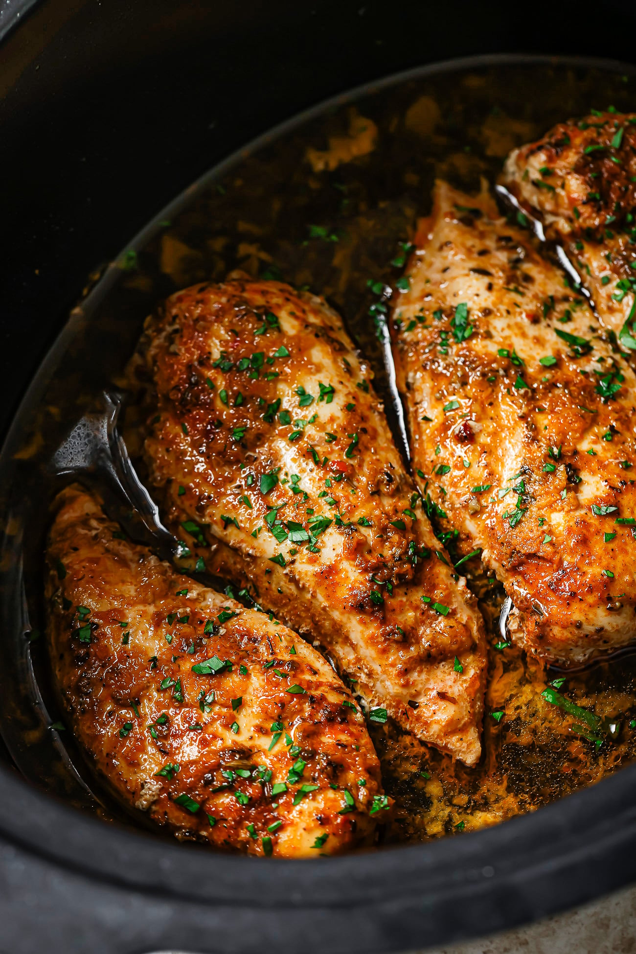 Three seasoned Slow Cooker Chicken Breasts sit in a dark slow cooker with flavorful sauce and sprinkled fresh herbs, showcasing a juicy and tender appearance.