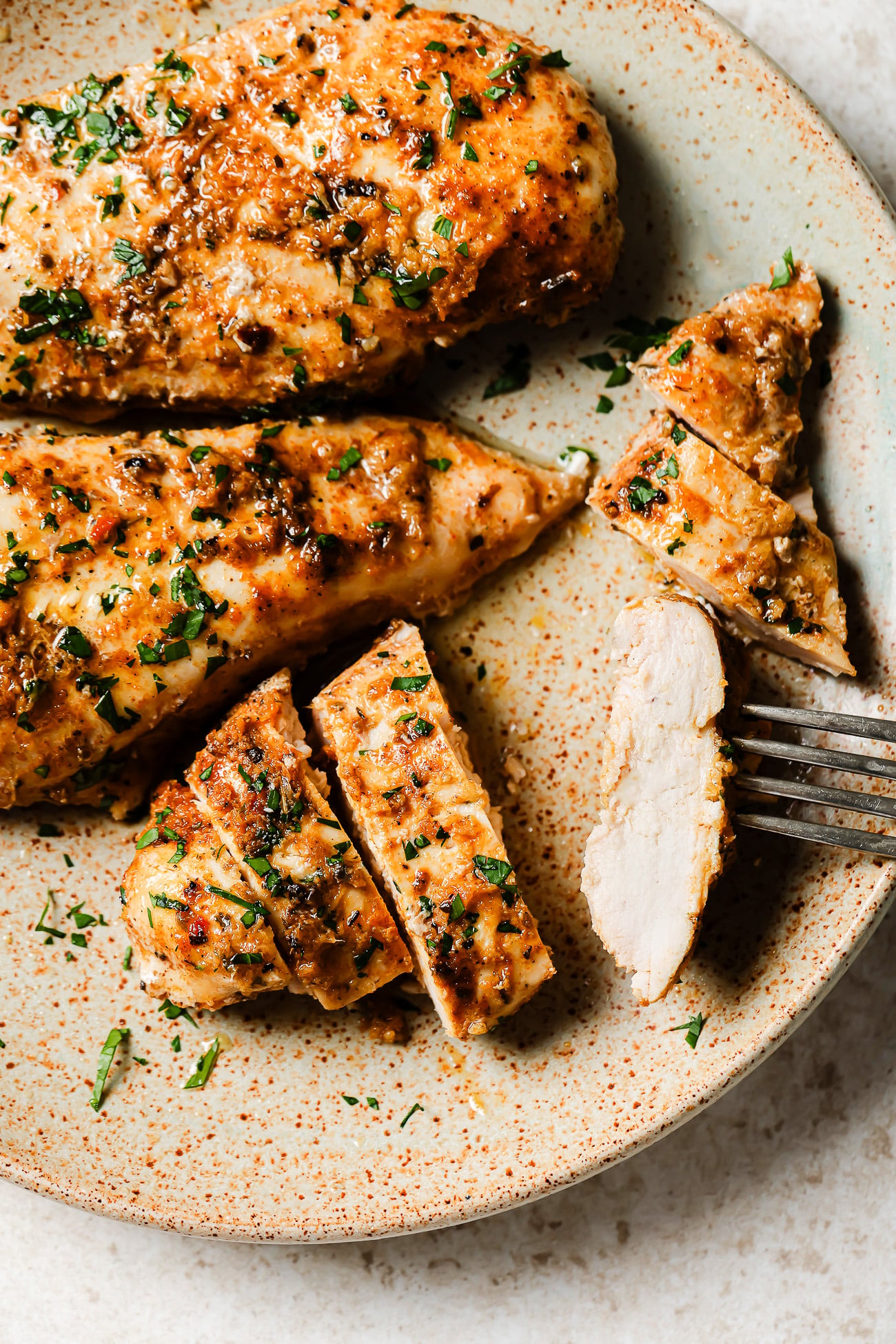 A plate with cooked, seasoned Slow Cooker Chicken Breast garnished with chopped herbs. One chicken breast is sliced, and a fork is holding a piece. The food is presented on a rustic, speckled plate.