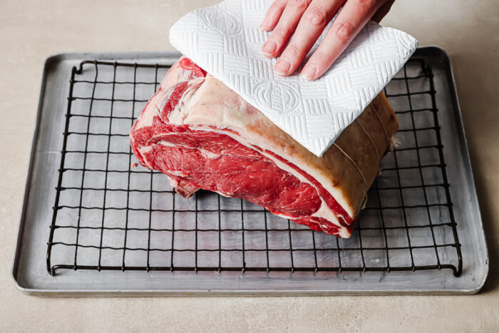 Patting dry a raw prime rib roast with a paper towel on a wire rack to remove moisture before roasting for the reverse sear prime rib.