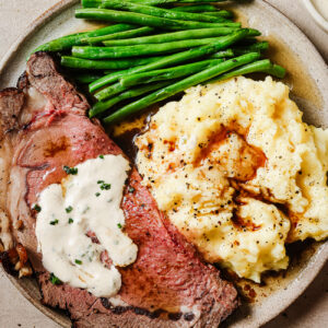 Juicy slices of reverse sear prime rib served with mashed potatoes, green beans, and horseradish sauce on a plate. A perfect holiday dinner.