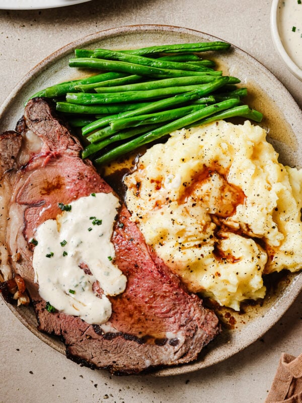 Juicy slices of reverse sear prime rib served with mashed potatoes, green beans, and horseradish sauce on a plate. A perfect holiday dinner.